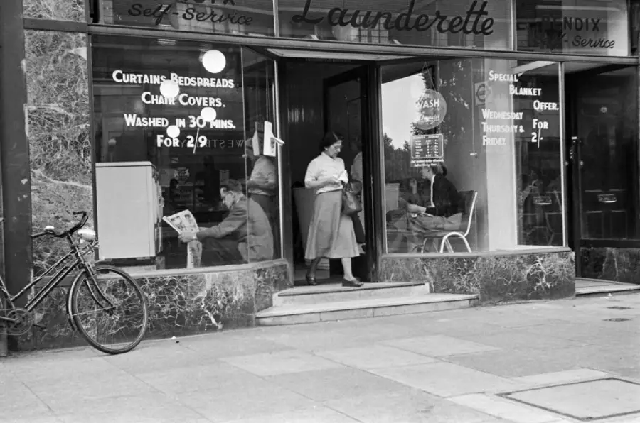 A woman exits a laundromat with glass windows; people inside sit and read. A bicycle leans against the building. Signs in the window advertise laundry services and prices.