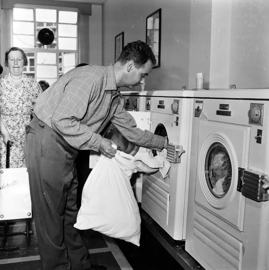 A man loads laundry into a washing machine at a laundromat while a woman waits nearby, holding a laundry basket.