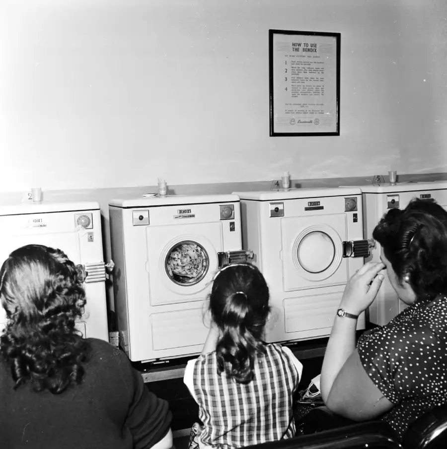 Three people, seen from behind, sit on a bench watching clothes spin in a row of washing machines in a laundromat. A framed instruction sign hangs on the wall above the machines.