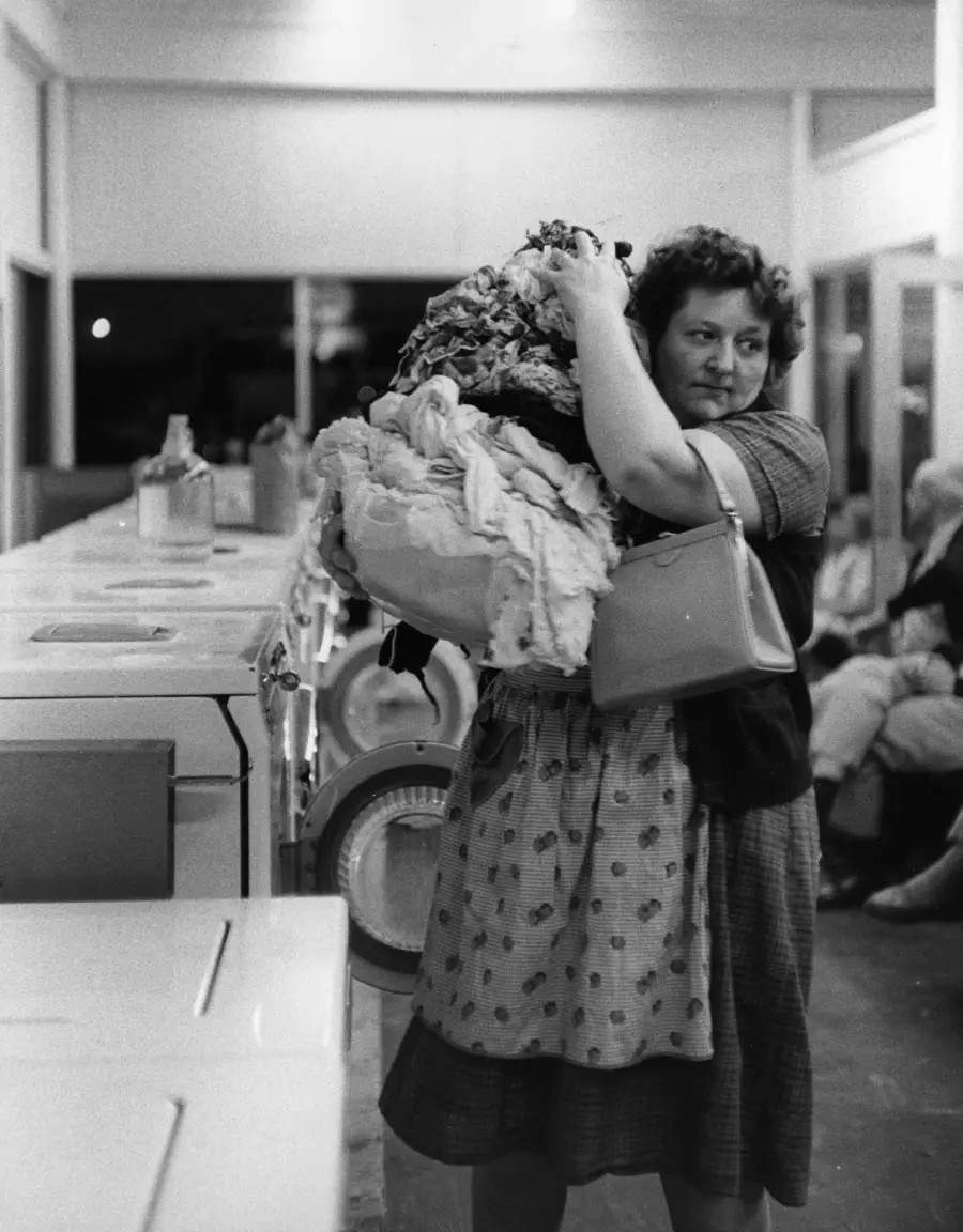 A woman in a laundromat carries a large pile of laundry and a purse, looking tired. Washing machines line the wall, and people sit on benches in the background. Black and white photo.