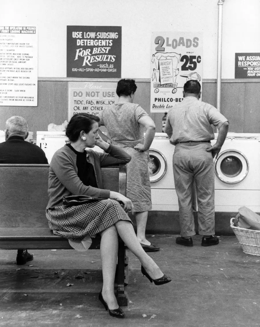 A woman sits on a bench in a laundromat, resting her head on her hand. Two people stand at washing machines. Signs about detergents and laundry prices hang on the wall.