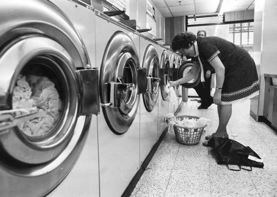 A woman loads clothes into a row of large washing machines at a laundromat, with a laundry basket and a coat on the floor beside her. A man stands in the background.