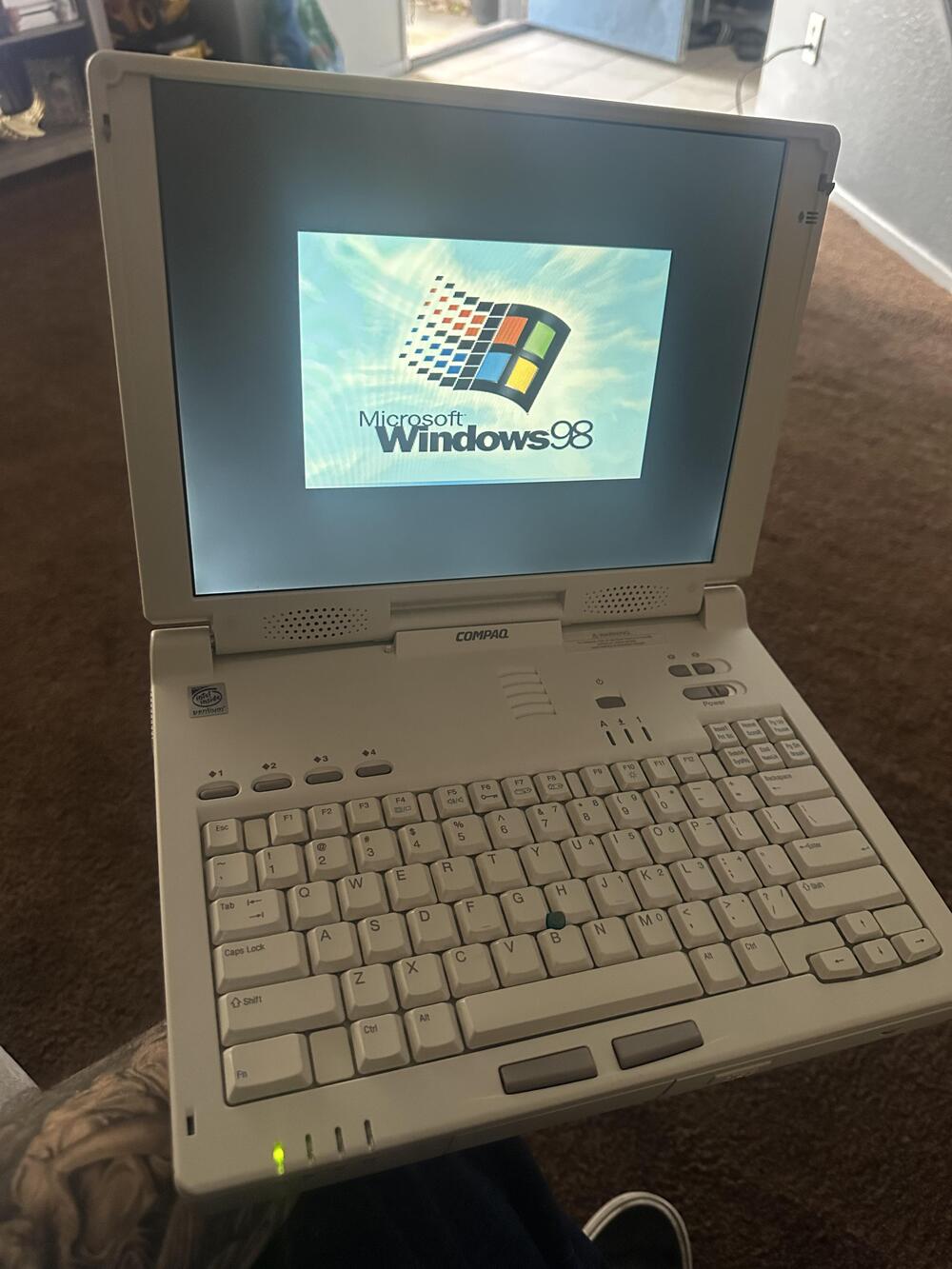 A person holds a vintage Compaq laptop displaying the Windows 98 startup screen in a home setting.
