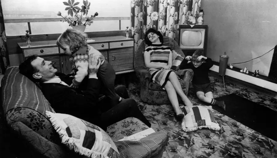A family relaxes in a living room: a man plays with a small child on an armchair, a woman reclines on a sofa, and another child sits on the floor near a television and patterned curtains.