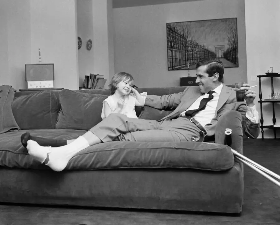 A man in a suit lounges on a sofa with a smiling young girl. He has one leg bandaged and crutches nearby. Both appear happy and relaxed in a modern living room.
