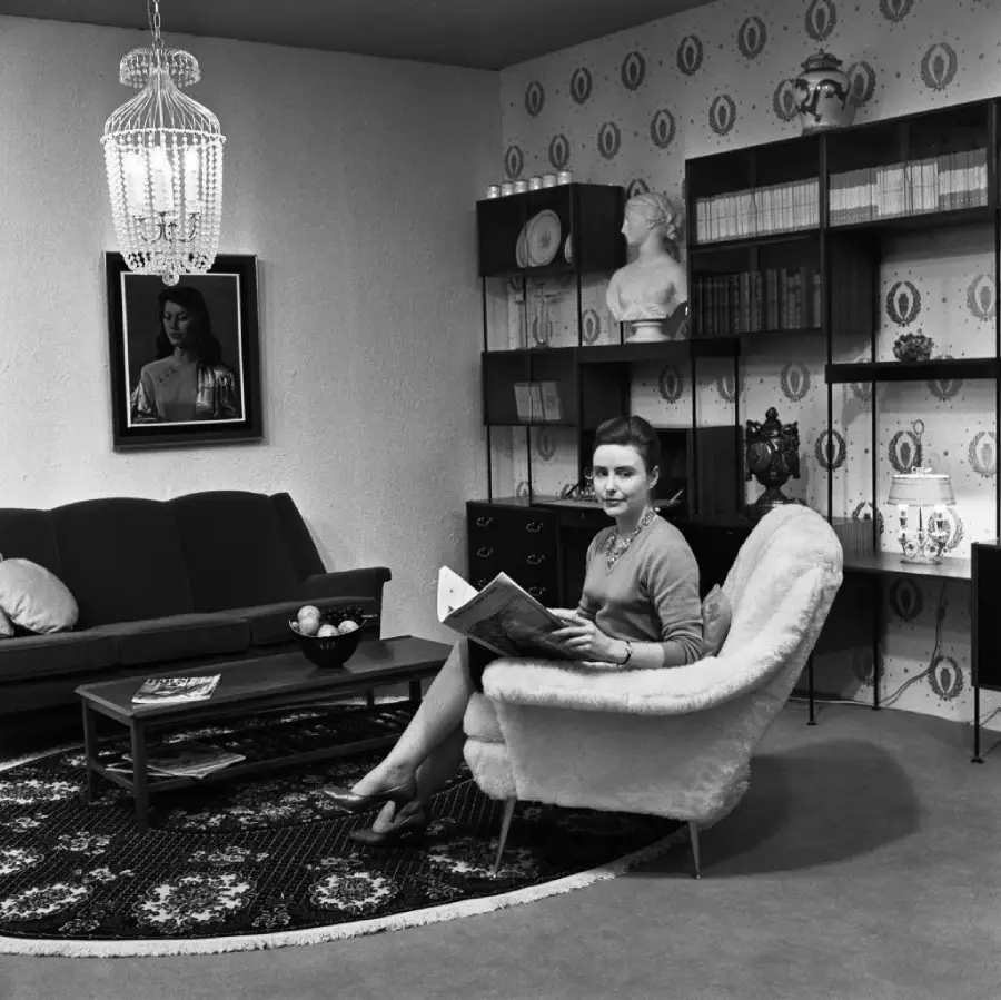 A woman sits in a cozy armchair reading a magazine in a stylish, mid-century living room with patterned wallpaper, shelves, a sofa, a coffee table, and a chandelier overhead.