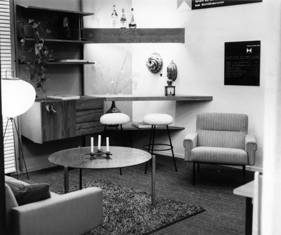 A mid-century modern living room with armchairs, a round coffee table with candles, floating shelves with bottles and decor, and a shag rug on the floor.