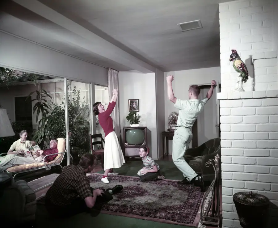 A family in a living room: two adults dance, one boy sits on the floor, another adult lounges on a couch, and a woman sits by the glass doors. A vintage TV and houseplants are in the background.