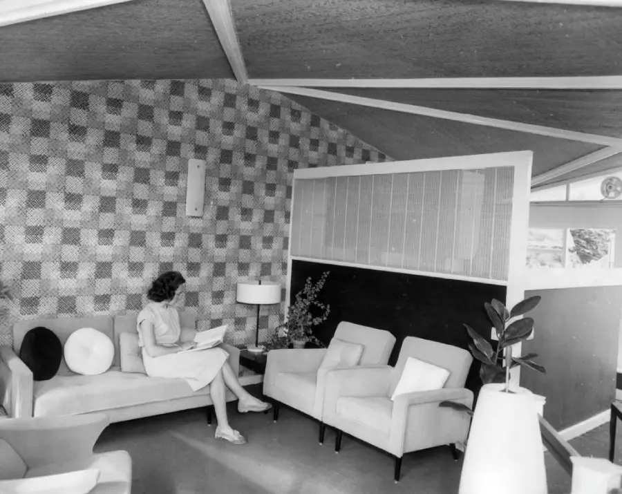 A woman sits on a sofa reading in a mid-century modern living room with patterned walls, armchairs, a lamp, and a large potted plant.