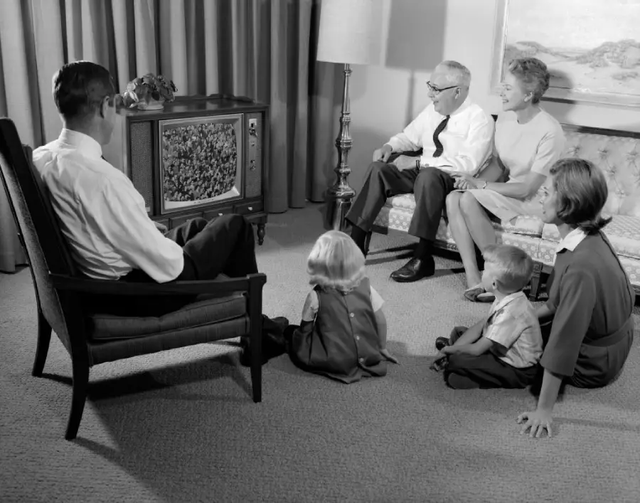 A family sits together in a living room, watching TV. Two adults sit on a couch, one in a chair, and three young children sit on the floor. The image is in black and white.