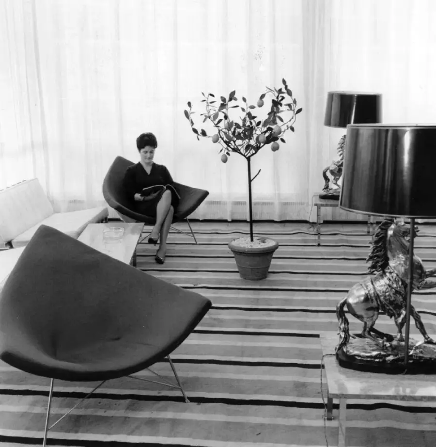 A woman sits reading in a modern living room with cone chairs, striped rug, potted plant, and metallic horse lamp. White curtains cover large windows in the background.