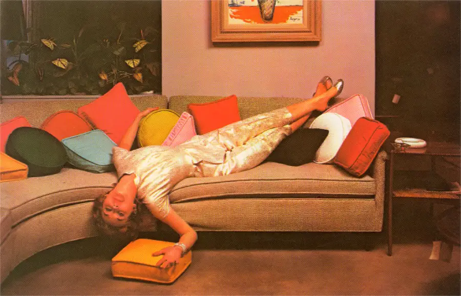 A woman in a shiny outfit lounges upside down on a curved sofa covered in colorful pillows, holding a yellow cushion and resting her feet on the sofa’s back. A painting and plants are visible in the background.