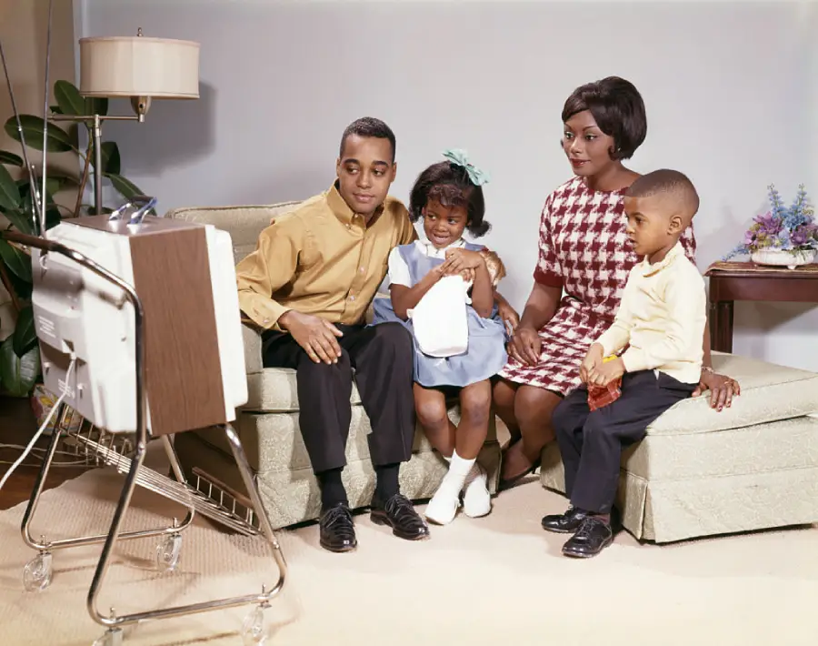 A family of four, dressed in 1960s attire, sits on a sofa watching television together in a living room setting. The parents and two children appear engaged and happy.