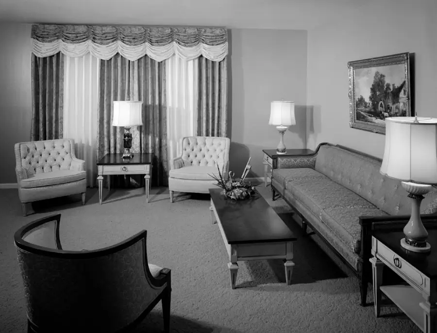 Black-and-white photo of a vintage living room with two armchairs, a sofa, a coffee table, two side tables with lamps, a curtained window, and a framed painting on the wall.