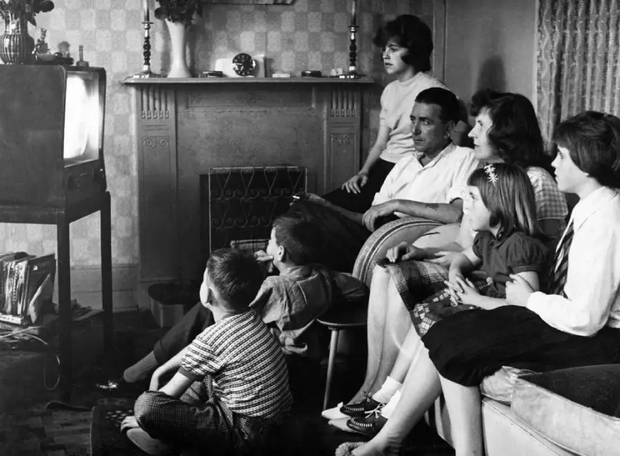 A black-and-white photo shows a family of seven sitting together in a living room, watching television. The children are sitting on the floor and sofa, while adults sit together on a couch, all focused on the TV.