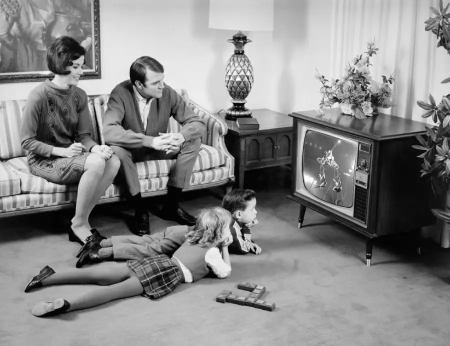 A family in 1960s attire sits in a living room watching television; parents on a sofa, daughter lying on the floor, and a young boy sits with a dog, all focused on the TV screen.