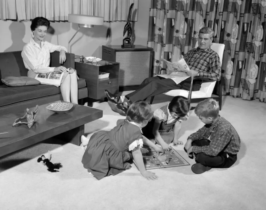 A woman sits on a couch reading, while a man relaxes in a chair with a newspaper. Three children play a board game on the carpeted floor in a cozy, mid-century living room.