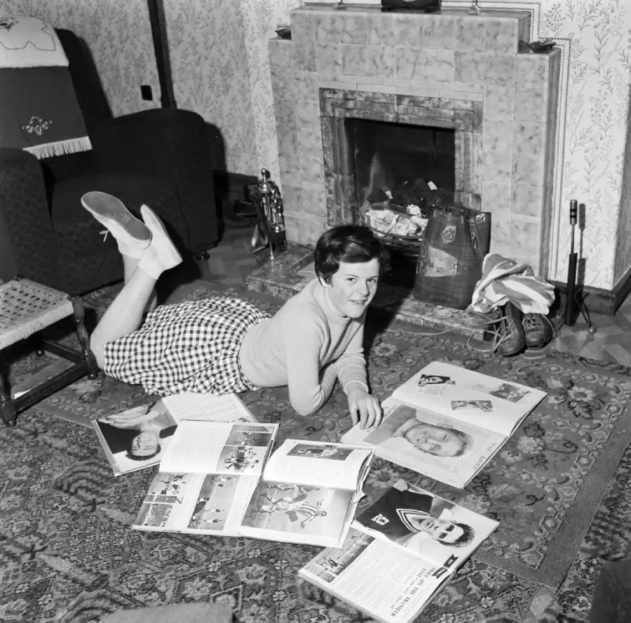 A woman lies on her stomach on a rug, smiling at the camera, surrounded by open magazines in a cozy living room with a fireplace and patterned wallpaper.