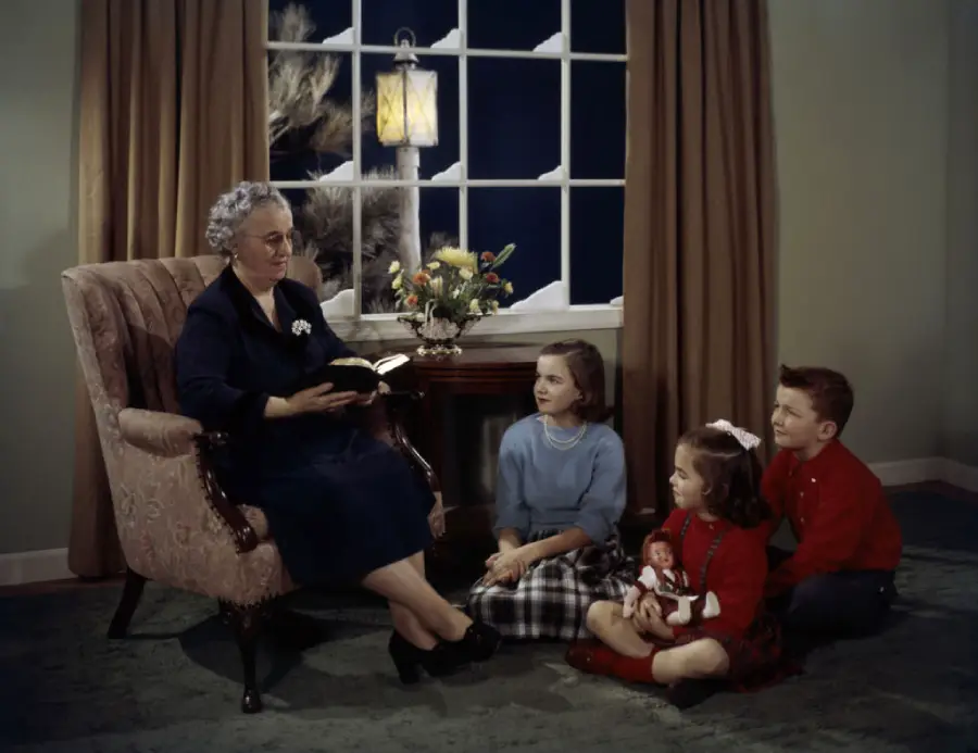 An elderly woman reads a book to three children sitting on the floor in a cozy living room, with a window, lamp, and flowers in the background. One child holds a doll.