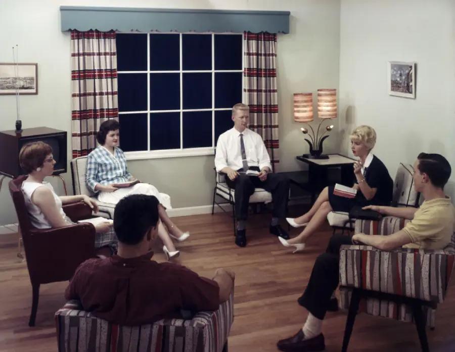 Six adults sit in a circle in a mid-century living room, engaged in conversation. The room has wood floors, striped curtains, patterned chairs, and two lamps on a side table by the window.