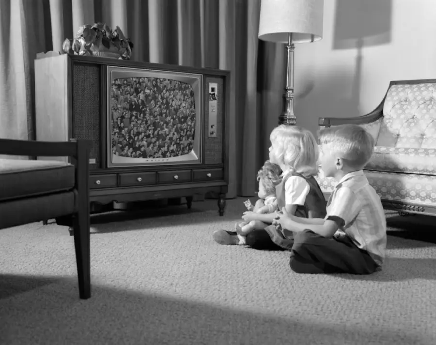 Two young children sitting on the carpet in front of an old-fashioned TV, watching a program. There’s a lamp, a sofa, and a chair in the living room. The image is black and white.