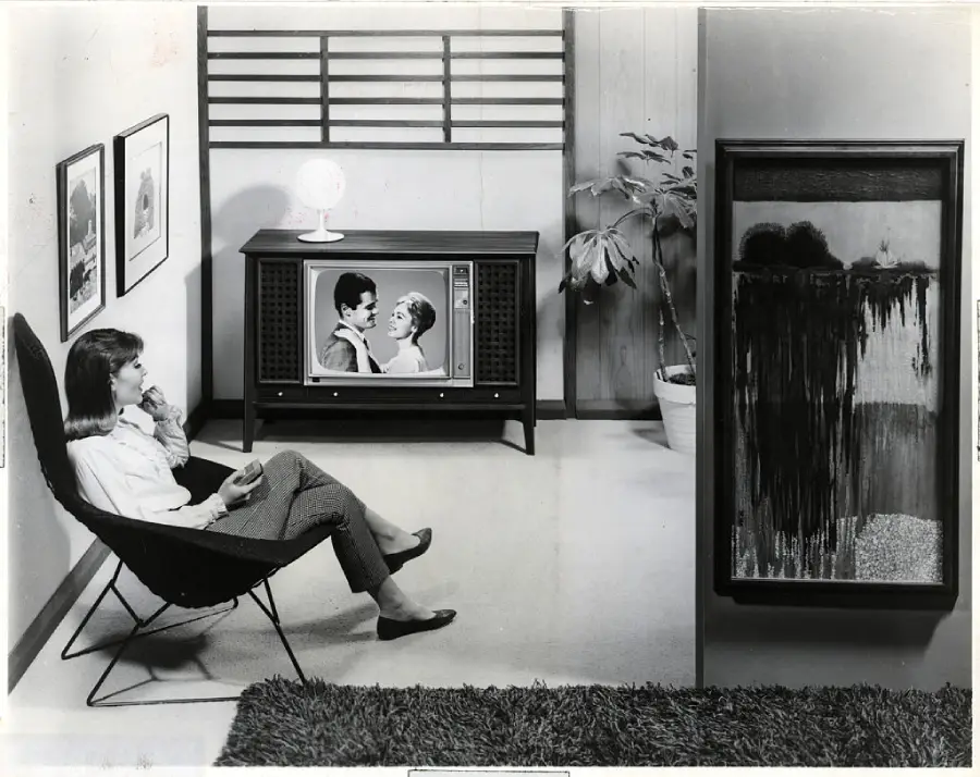 A woman sits in a modern chair, watching a black-and-white television featuring two people. The mid-century room has framed art, a potted plant, and a textured rug.
