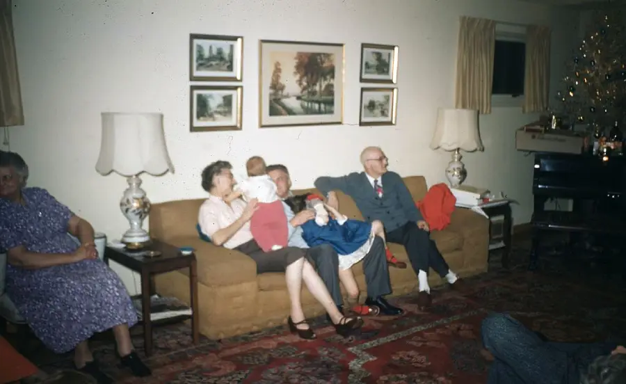 Five adults and two children sit on a brown couch in a living room with a decorated Christmas tree, lamps, and framed pictures on the wall. The atmosphere appears warm and festive.