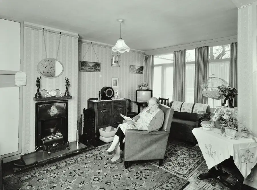 An elderly woman sits reading in an armchair in a cozy, vintage living room with patterned wallpaper, a fireplace, TV, and various decorations and furniture.
