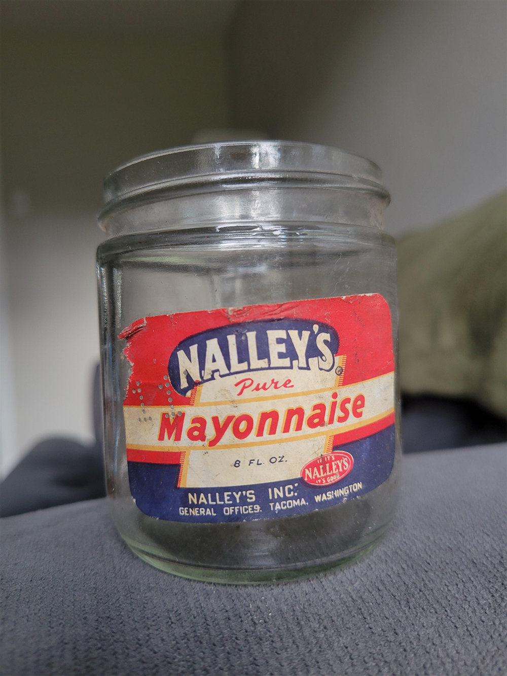 An empty vintage glass jar with a red, white, and blue Nalley’s Pure Mayonnaise label, sitting on a gray surface with a blurred background.