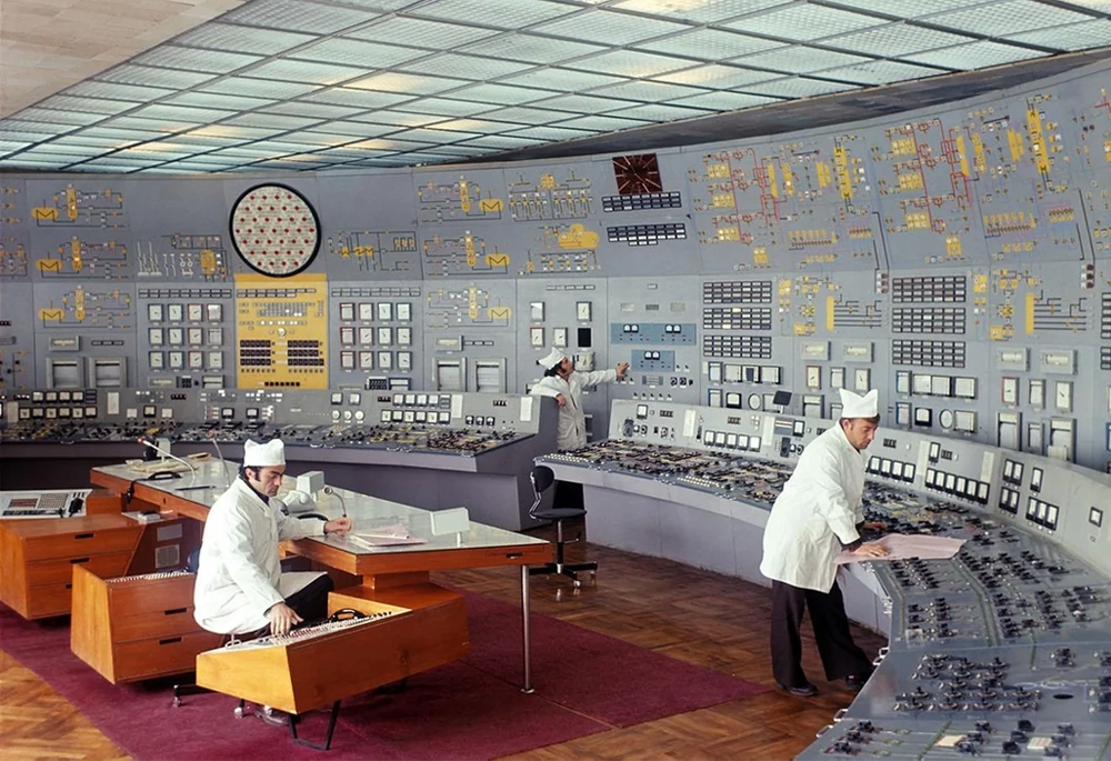 Three people in white lab coats and hats work at large, curved control panels filled with buttons, switches, and gauges in a retro control room with a high ceiling.