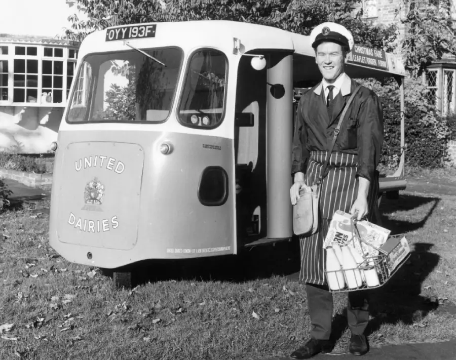 A smiling milkman in uniform stands beside a vintage United Dairies milk float, holding a basket with milk bottles and groceries. The scene appears to be set in a residential neighborhood.