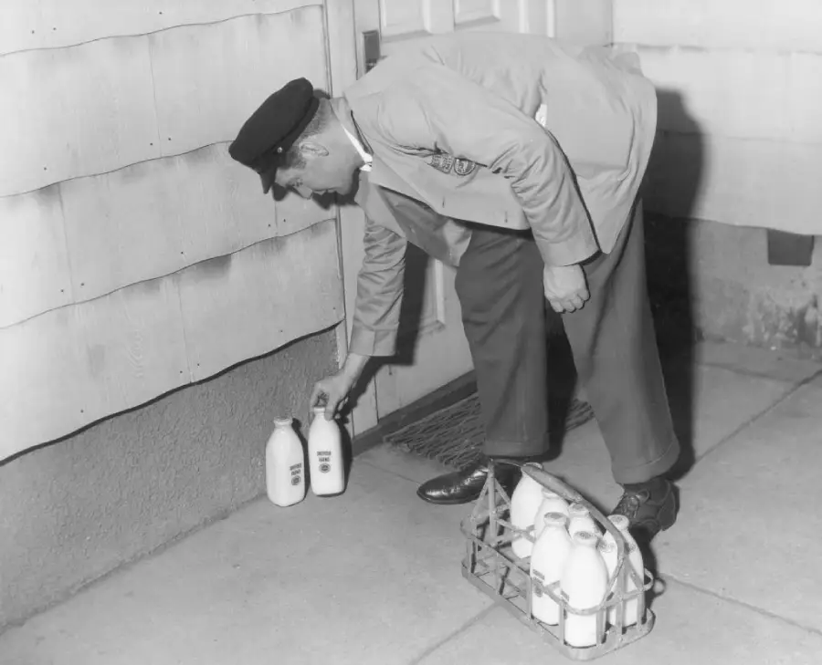 A milkman wearing a uniform and cap delivers glass bottles of milk to a doorstep, picking up old bottles while placing new ones from a metal carrier.