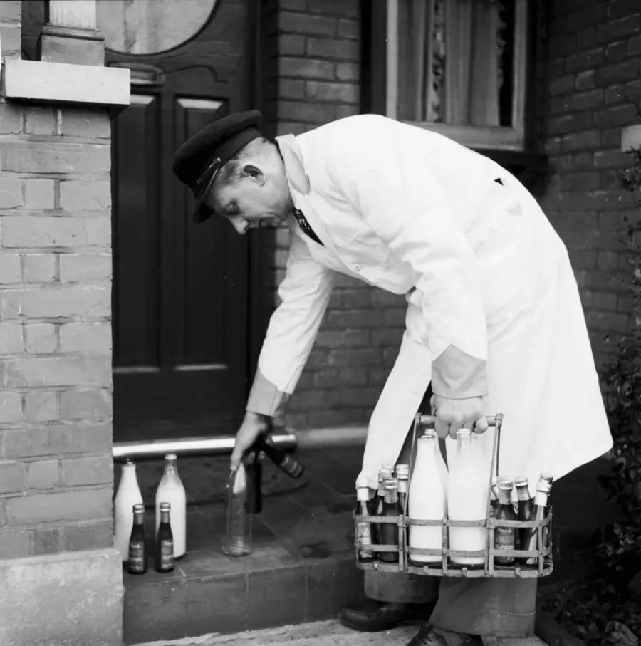 A milkman in a white coat and hat delivers glass bottles of milk to a doorstep, carrying a metal crate filled with bottles.