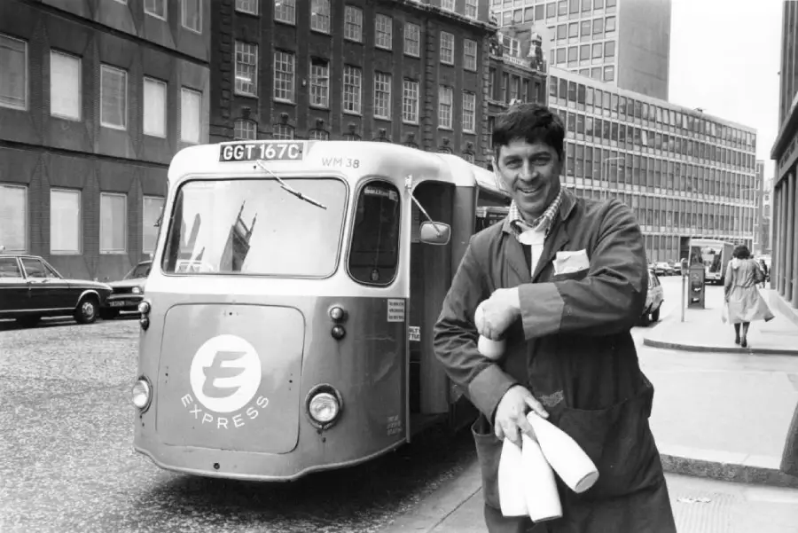 A smiling man in a work coat holds bottles on a city street beside a small vintage delivery van labeled “Express.” Buildings and pedestrians are visible in the background.