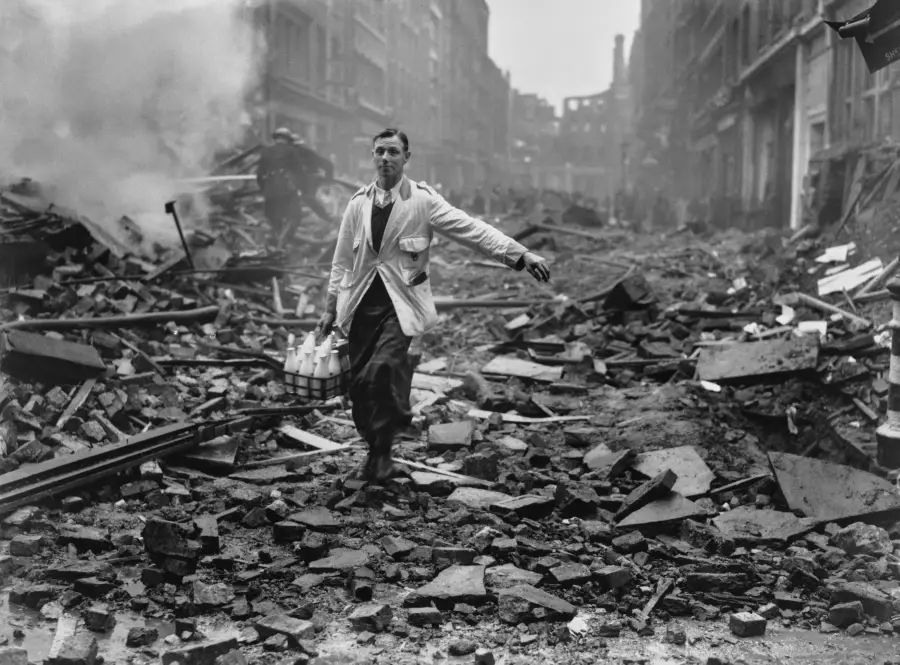 A milkman in a white coat carries bottles through rubble and debris on a destroyed city street, with smoke rising in the background and damaged buildings on both sides.