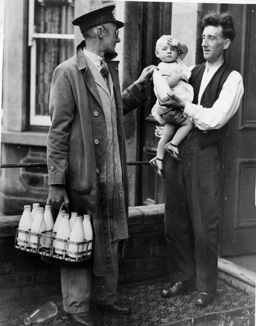 A milkman holding a crate of milk bottles talks to a man holding a baby outside a house. The baby is playfully saluting or shielding its eyes, while the men smile and converse near the front door.