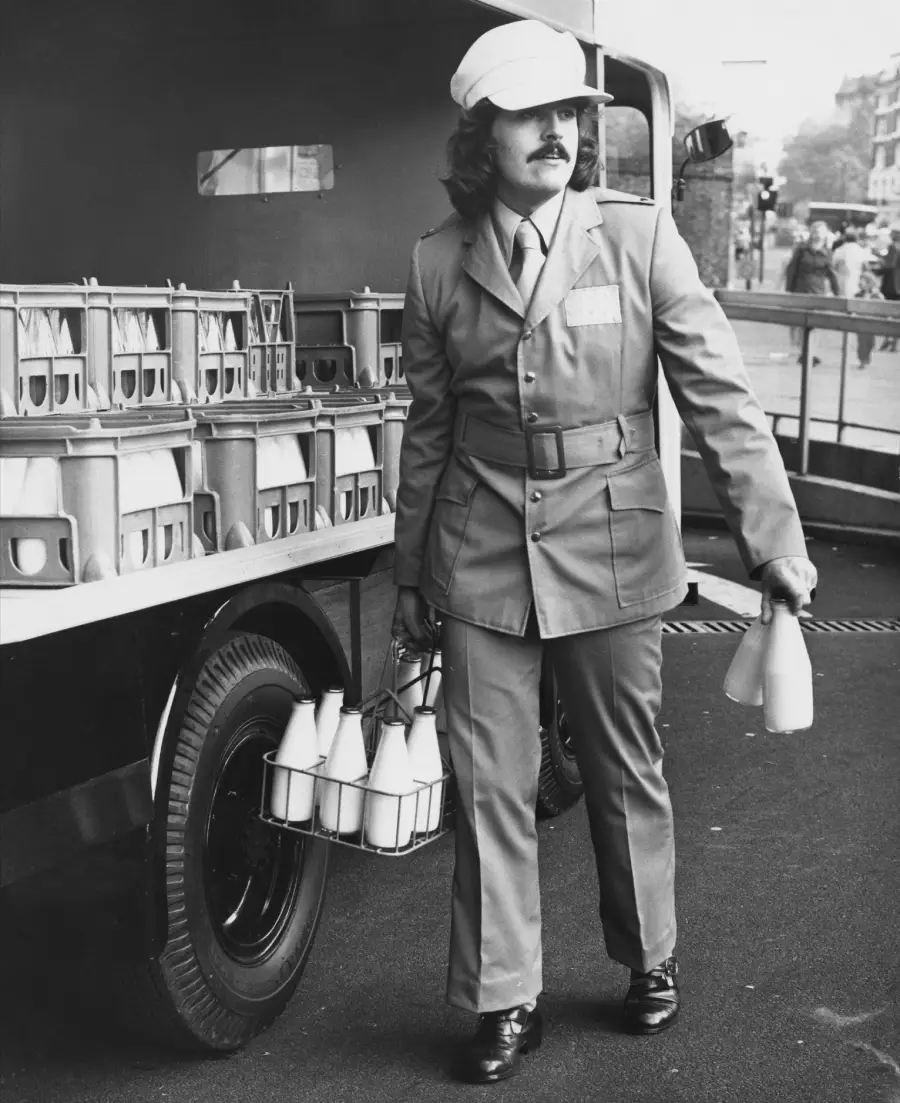 A milkman in uniform and cap carries a wire basket of glass milk bottles from a delivery truck parked on the street.