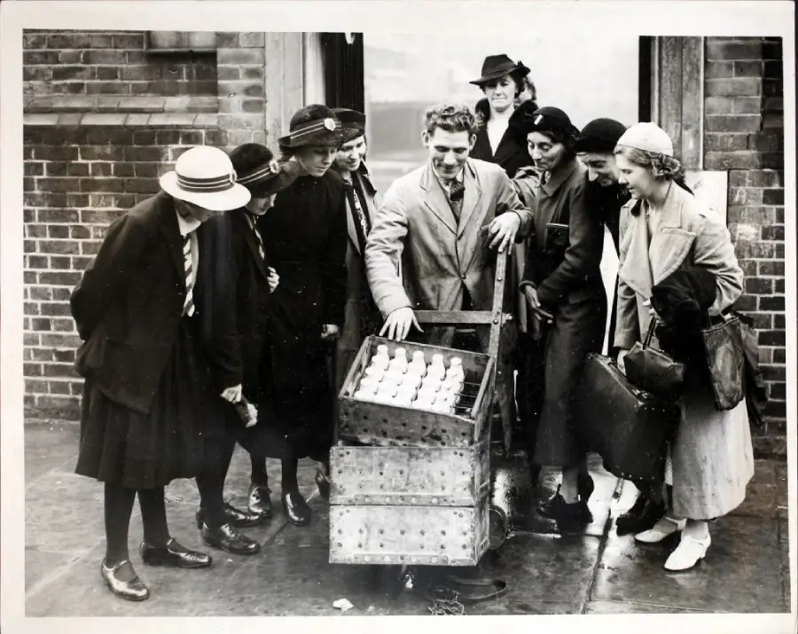 A group of people in vintage clothing gather around a man showing eggs in a crate on a sidewalk, with brick buildings in the background.