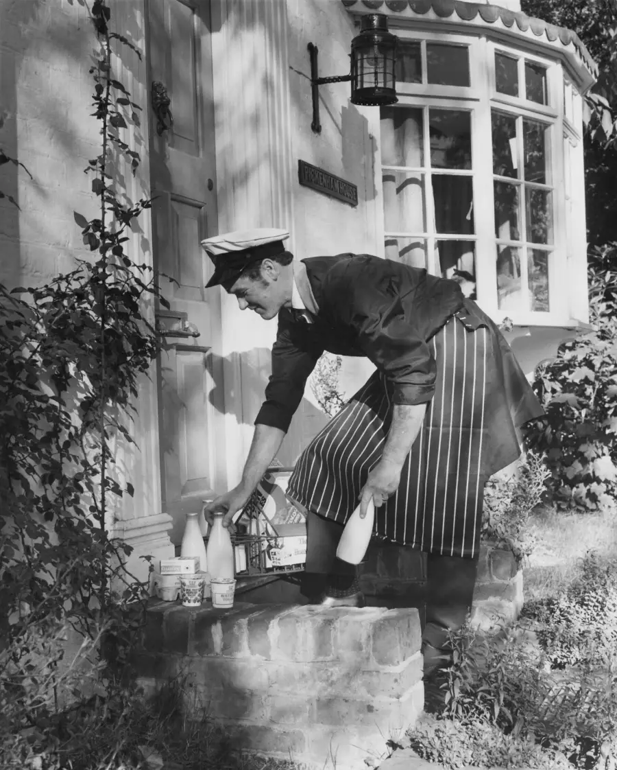 A milkman in uniform and striped apron delivers milk bottles and dairy products to the doorstep of a house with a rounded bay window.