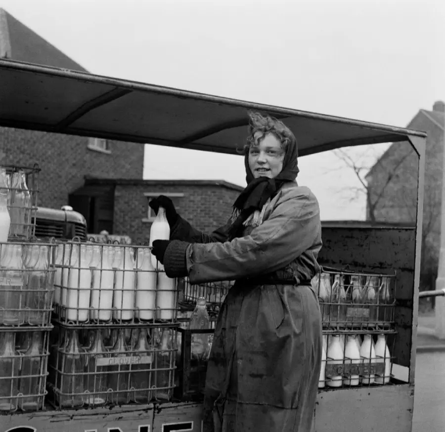A woman dressed in a coat and scarf smiles while delivering glass bottles of milk from a truck, with crates of milk bottles stacked behind her.