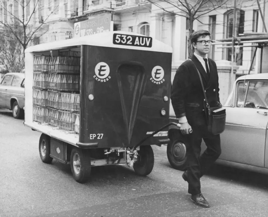 A man in a suit walks next to a small electric delivery vehicle with racks inside, labeled “532 AUV” and “Express,” on a city street lined with cars and buildings.