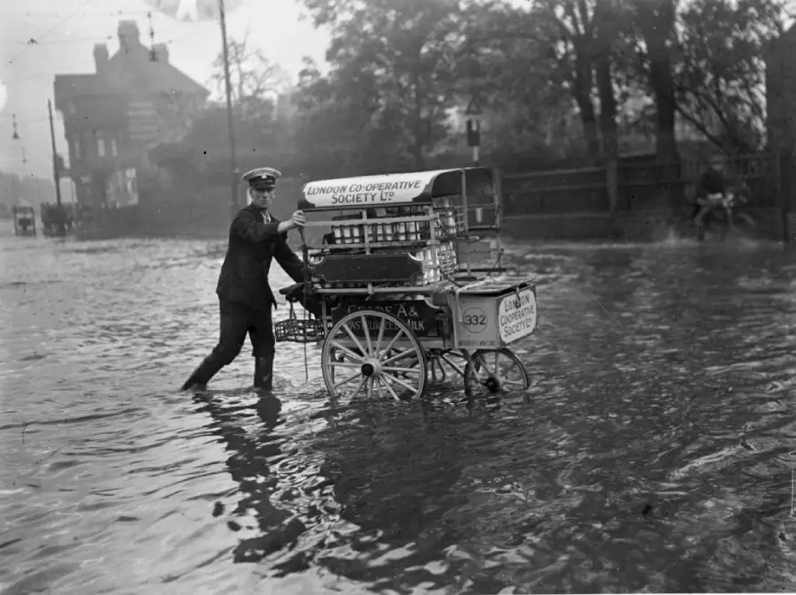 A man in uniform pushes a delivery cart labeled “London Co-Operative Society Ltd” through a flooded street, with water reaching above his ankles. Trees and buildings are visible in the background.