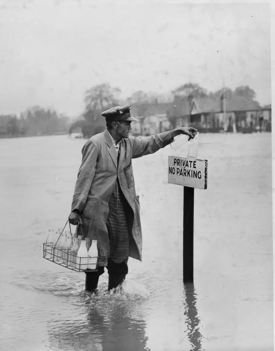 A milkman in uniform wades through floodwater, carrying a metal crate of milk bottles and resting his hand on a “Private No Parking” sign. Houses and trees are visible in the background.