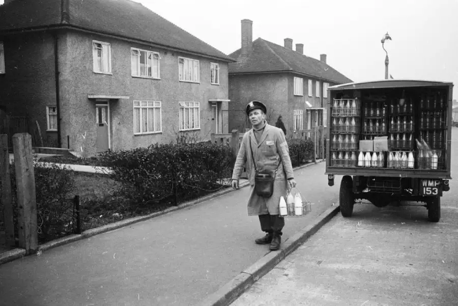 A milkman in uniform carries glass milk bottles from a delivery truck parked on a suburban street lined with houses, likely in the mid-20th century.