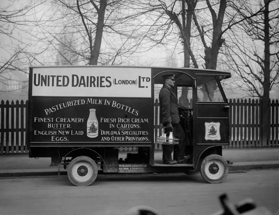A vintage United Dairies delivery van is parked, with a man holding a milk carrier standing at its open door. The van advertises milk, butter, eggs, and cream, with trees and a fence in the background.