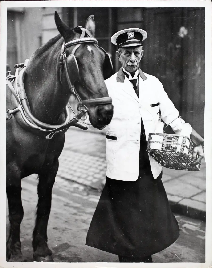 A stern-faced postal worker in uniform stands beside a horse on a cobblestone street, holding a wicker basket. The horse is harnessed and looking at the camera.