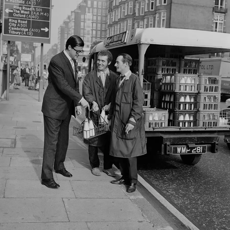 Three men stand on a city sidewalk by a milk delivery truck. One man in a suit receives a milk bottle crate from two milkmen in uniforms. Buildings and street signs are visible in the background.