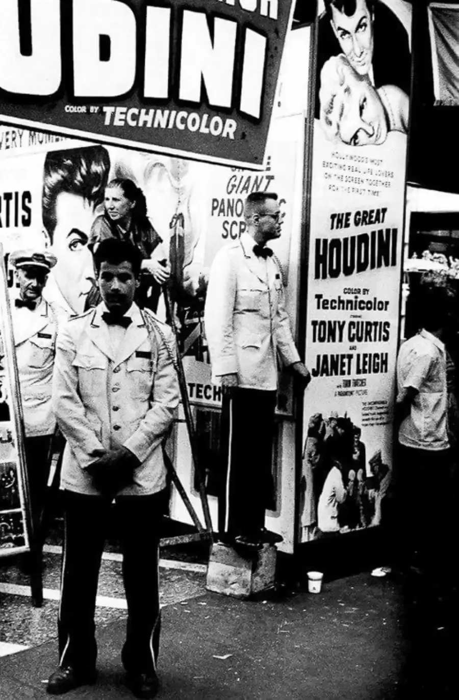 A black-and-white photo shows men in uniforms standing outside a theater displaying large posters for “The Great Houdini” starring Tony Curtis and Janet Leigh. Crowds and promotional signs fill the busy street scene.