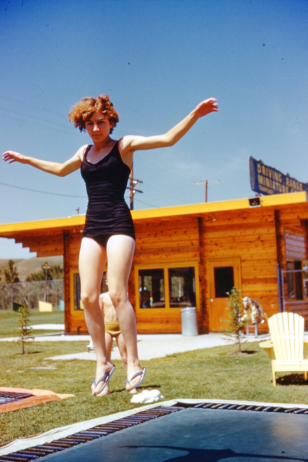 A woman in a black swimsuit and flip-flops jumps on a trampoline outside, with a wooden building and yellow chairs in the background on a sunny day.