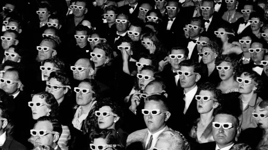 A large audience sits closely together, all wearing white 3D glasses and watching something intently. The image is black and white, and everyone appears focused and engaged.